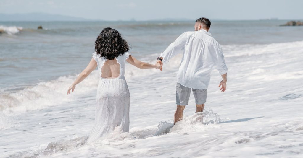 A couple in summer attire walks hand in hand along the beach, enjoying a sunny day.