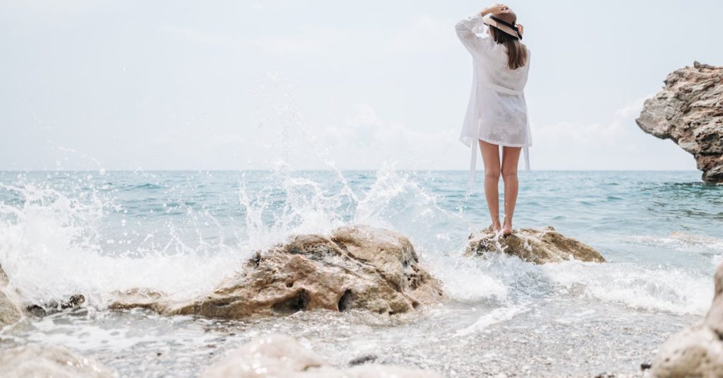 Woman on rocky shore with splashing waves, enjoying a sunny day at the beach.